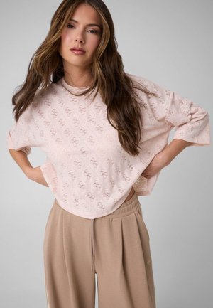 Jeune femme aux longs cheveux portant un haut ample léger rose clair à motifs et un pantalon beige à cordon, posant avec les mains sur les hanches devant un fond gris.