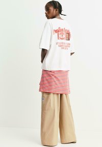 Person med fletninger i håret iført en hvid oversized T-shirt med rød tekst, en rød- og hvidstribet nederdel og beige bukser med brede ben, stående sidelæns.