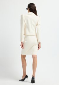 Creme blazer med skulderpuder, cropped design, lange ærmer, knapdetaljer, paired med en tætsiddende creme nederdel og sorte højhælede sko.