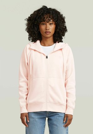 Woman with curly hair wearing a light pink zip-up hoodie over a white shirt and blue jeans, standing against a plain background.