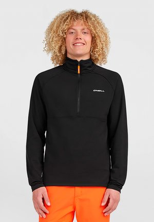 Young person with curly hair wearing a black O'Neill zip-up top and bright orange pants, standing and smiling against a plain background.