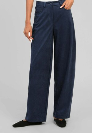 Personne portant un pantalon large bleu foncé avec des poches avant et des chaussures plates noires, debout devant un fond clair uni.