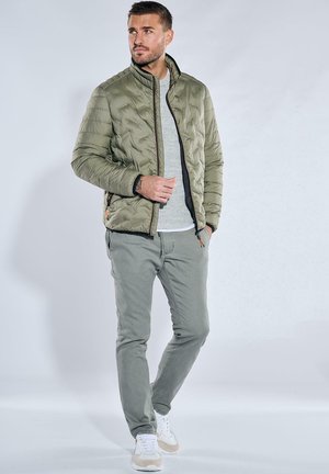 Olive green puffer jacket with quilted texture, black zipper, worn over a light gray sweater and gray pants, paired with white sneakers.