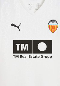 Camiseta deportiva blanca con un patrón texturizado. Presenta el logo negro de Puma y el emblema del Valencia CF, además de un logo negro del TM Real Estate Group.