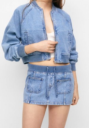 Kvinde iført lyseblå denimjakke med lynlås og matchende kort nederdel med store forlommer, der holder i lynlåsen på jakken.