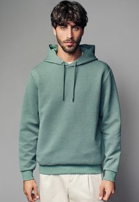 Sudadera de color verde menta texturizada con capucha ajustable, puños y dobladillo acanalados, que presenta un patrón geométrico en toda la tela.