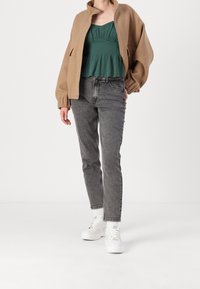 Giacca oversize color cammello, top verde con arricciatura, jeans grigi e sneakers bianche. La giacca ha un colletto rialzato; il top presenta un orlo arricciato.