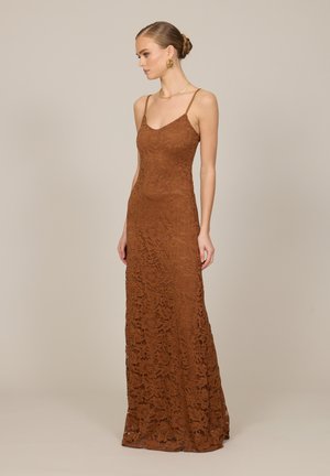 Femme aux cheveux en chignon portant une longue robe ajustée en dentelle marron à fines bretelles, debout de profil sur un fond beige uni.