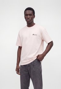 Giovane uomo che indossa una t-shirt rosa chiaro con la scritta "Dropsize" e pantaloni grigi, posando con una mano in tasca su uno sfondo semplice.