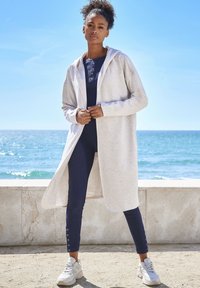 Cárdigan con capucha de color gris claro sobre un atuendo azul marino con detalles de botones. Combinado con zapatillas deportivas grises en un fondo junto a la playa.