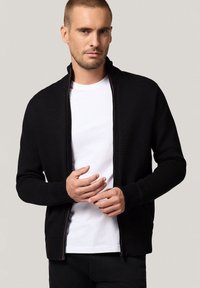 Czarna bluza na zamek z prążkowanymi mankietami i dołem, z wysokim kołnierzem i teksturowanym wzorem, nałożona na białą koszulkę.