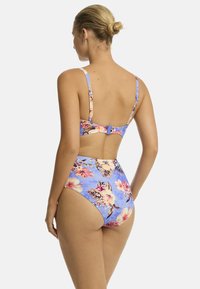 Een vrouw met een knot draagt een blauwe bloemenbikini van achteren, waarbij ze high-waisted bottoms en een strappy bikini top laat zien tegen een effen achtergrond.