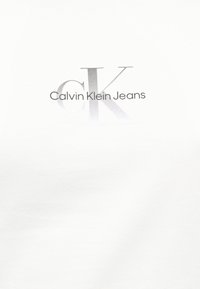Calvin Klein Jeans logó nagy, kifakult „CK” betűkkel a háttérben, és a márkanév tiszta szöveggel fehér anyagon.