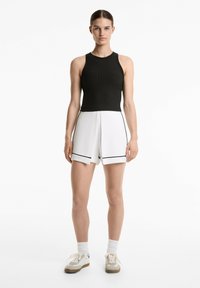 Svart ribbad linne tillsammans med vita shorts med svarta detaljer. Riven design vid fållen. Modell som bär beige sneakers och vita strumpor.