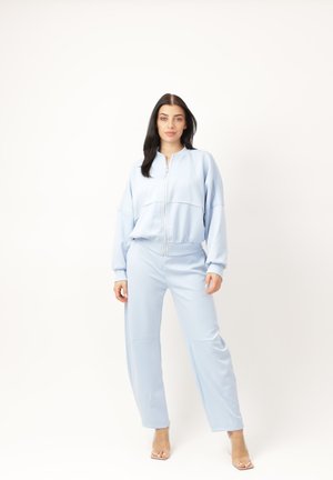 Mujer de pie contra un fondo blanco, lleva una chaqueta azul claro con cierre y pantalones sueltos a juego, además de zapatos de tacón alto transparentes.