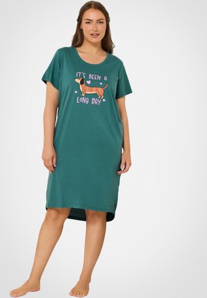 Vrouw draagt een teal knielange jurk met een teckel en de tekst "Het is een lange dag geweest" op de voorkant, staat op blote voeten.