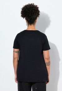 Camiseta negra de manga corta con cuello redondo. Tejido de algodón suave, corte relajado y dobladillo trasero un poco más largo. Tatuaje visible en el brazo.