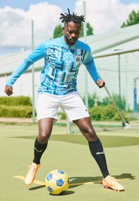 Maglietta da calcio a maniche lunghe con motivo blu e bianco, pantaloni corti bianchi, calze nere e scarpini arancioni. Giocatore che dribbla un pallone da calcio giallo su un campo verde.