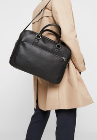 Schwarze Lederhandtasche mit strukturierter Oberfläche, ausgestattet mit zwei oberen Griffen und einem verstellbaren Schultergurt. Seitliche Reißverschlusstasche für zusätzlichen Stauraum.