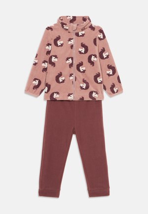 Fleece pižamos komplektas, sudarytas iš rožinės užtraukiamos viršutinės dalies su baltais ir rudais vienaragio raštais, derinamas su vientisomis tamsiai raudonomis kelnėmis. Minkšta tekstūra, elastingos rankogaliai.