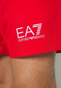 Červené plavky s logom "EA7" a textom "Emporio Armani". Materiál vyzerá hladko a dizajn je jednoduchý a funkčný.