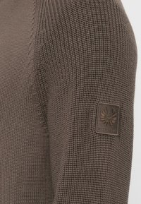 Ruskea neulepuseron hiha, jossa on tekstuurinen alue, jossa on siipisymboli ja "BELSTAFF"-teksti ylävarressa.