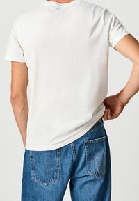 Mand iført en ensfarvet hvid kortærmet T-shirt og blå denimjeans, hånd der trækker lidt i T-shirten bagpå.