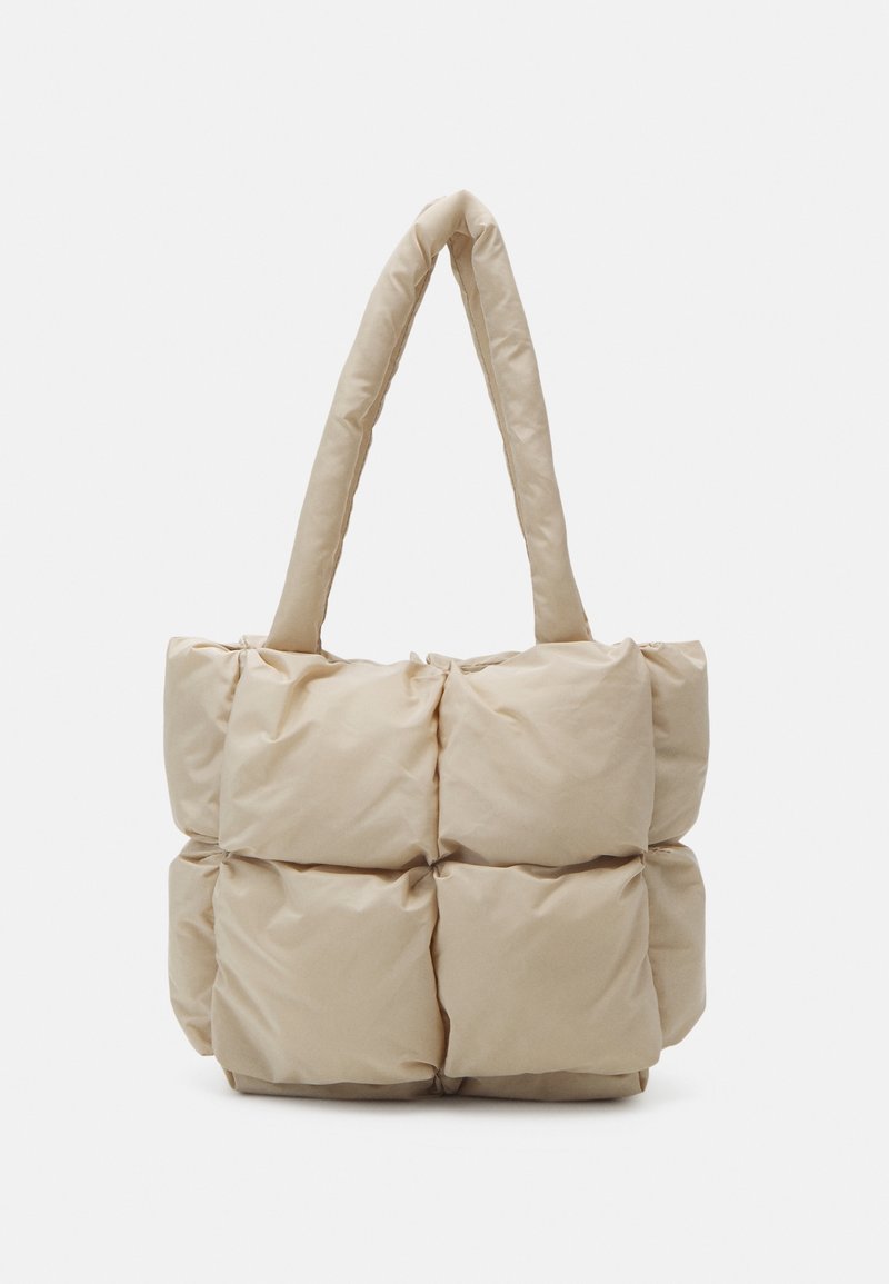 Sac fourre-tout matelassé beige avec deux poignées rembourrées, présentant une surface douce et texturée ainsi qu'une forme carrée avec des carrés cousus visibles.