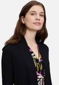 Een vrouw met schouderlang donker haar glimlacht, gekleed in een zwarte blazer over een kleurrijke bloemenblouse. Eenvoudige witte achtergrond.