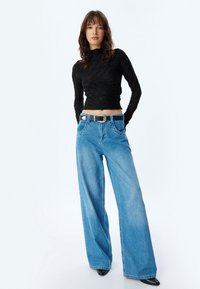 Schwarzes, strukturiertes Langarmoberteil, kombiniert mit weit geschnittenen blauen Jeans. Besitzt einen schwarzen Gürtel mit einer silbernen Schnalle.