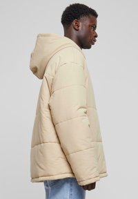 Beige pufferjacka med huva, med en kviltad design och synlig sömnad. Mjuk textur, något oversized passform med en fållad nederkant.