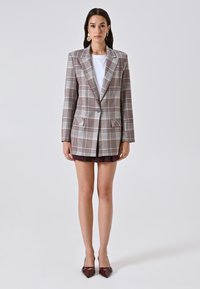 Geruite blazer met een lichtgrijze, bruine en bordeauxrode print. Enkele knoopsluiting, twee voorzakken en een getailleerde pasvorm. Wit shirt eronder.