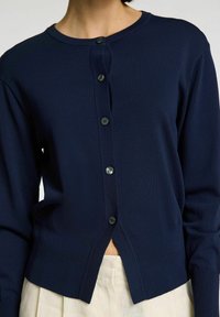 Námořnická modrá pletená cardigan s kulatým výstřihem, předním zapínáním na pět tmavých knoflíků a mírně zkráceným střihem.