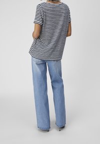 Person wearing a black and white striped short-sleeve shirt and light blue wide-leg jeans, standing with hands in pockets facing away.