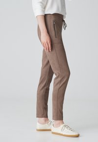 Braune, figurbetonte Hosen aus weichem, wildlederähnlichem Material. Verfügt über Reißverschluss-Seitentaschen und einen Kordelzugbund, kombiniert mit weißen Sneakers.