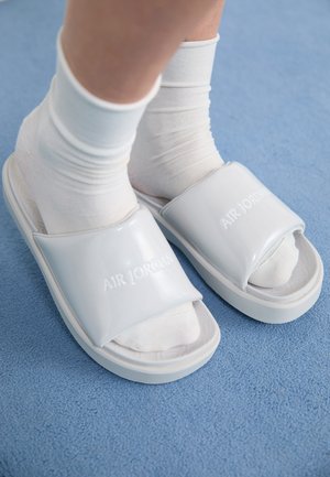 White Air Jordan slide sandals with a glossy finish, featuring an open-toe design and textured inner sole, worn over white socks.