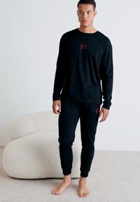 Chemise noire à manches longues avec un petit logo rouge sur la poitrine, assortie à un pantalon noir ajusté. Texture douce, coupe décontractée, pieds nus.