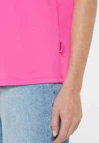 Nahaufnahme einer Person, die ein leuchtend pinkes Shirt mit dem Tag "SENSES" und hellblauen Jeans trägt, mit entspanntem Hand neben dem Körper.