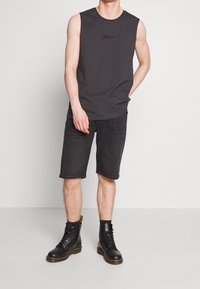 Man wearing sleeveless black shirt, black knee-length shorts, black socks, and black lace-up boots standing against white wall.