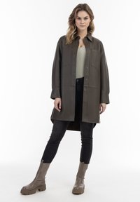 Olivengrøn faux læder oversized skjortejakke med krave, to brystlommer og trykknapper, parret med sorte jeans og chunky beige støvler.