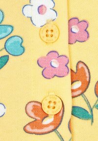 Keltainen neulevillatakki, jossa on värikkäitä kukkakuoseja vaaleanpunaisena, sinisenä ja punaisena. Kiinnitetty pyöreällä keltaisella napilla.