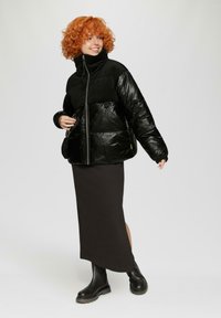 Zwarte pufferjas met een glanzende tekstuur, hoge kraag en ritssluiting. Draagt over een lange, bruine rok en zwarte chunky laarzen.