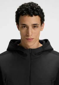 Svart huva jacka har en mjuk textur, framdragkedja och två fickor. Designen är elegant med minimala sömmar och en tight passform.