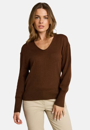 Femme aux cheveux châtain clair, portant un pull marron à manches longues et col en V, et un pantalon beige, debout devant un fond uni.