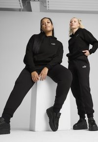 Schwarze Hoodies und Sweatpants aus weichem Stoff, mit einem kleinen Logo. Beide Personen tragen schwarze Plateausneaker. Einfacher Studiohintergrund.