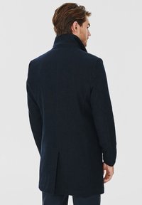 Granatowy wełniany płaszcz z wysokim kołnierzem i dopasowanym krojem, z jednym rozcięciem z tyłu i czystymi liniami w minimalistycznym stylu.