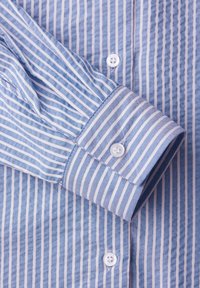 Blau-weiß gestreiftes Hemd mit langen Ärmeln und weißen Knöpfen, das einen hochgekrempelten Ärmel und eine geknöpfte Manschette zeigt.