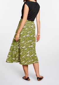 Green skirt with a sailboat pattern, flared design, mid-length, paired with a fitted black top, and worn with flat brown sandals.