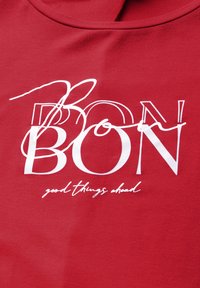 Camiseta roja con cuello redondo que presenta letras en negrita de color blanco que dicen "BON" y "buenas cosas por venir" en escritura cursiva. Textura de tela suave.