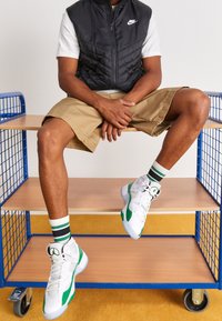 Svart puffig väst över en vit T-shirt, khakishorts, vita och gröna hög-top sneakers, randiga gröna och vita strumpor, sittande på en hylla.